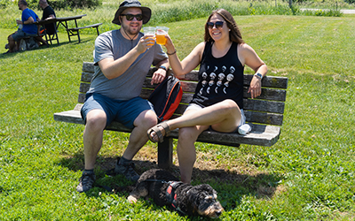 Two people on a bench holding beverages with a dog laying in the grass