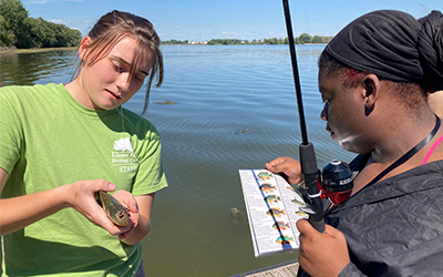 A staff member holding a fish and showing it to a person from the public holding a fishing pole and a fish ID chart.
